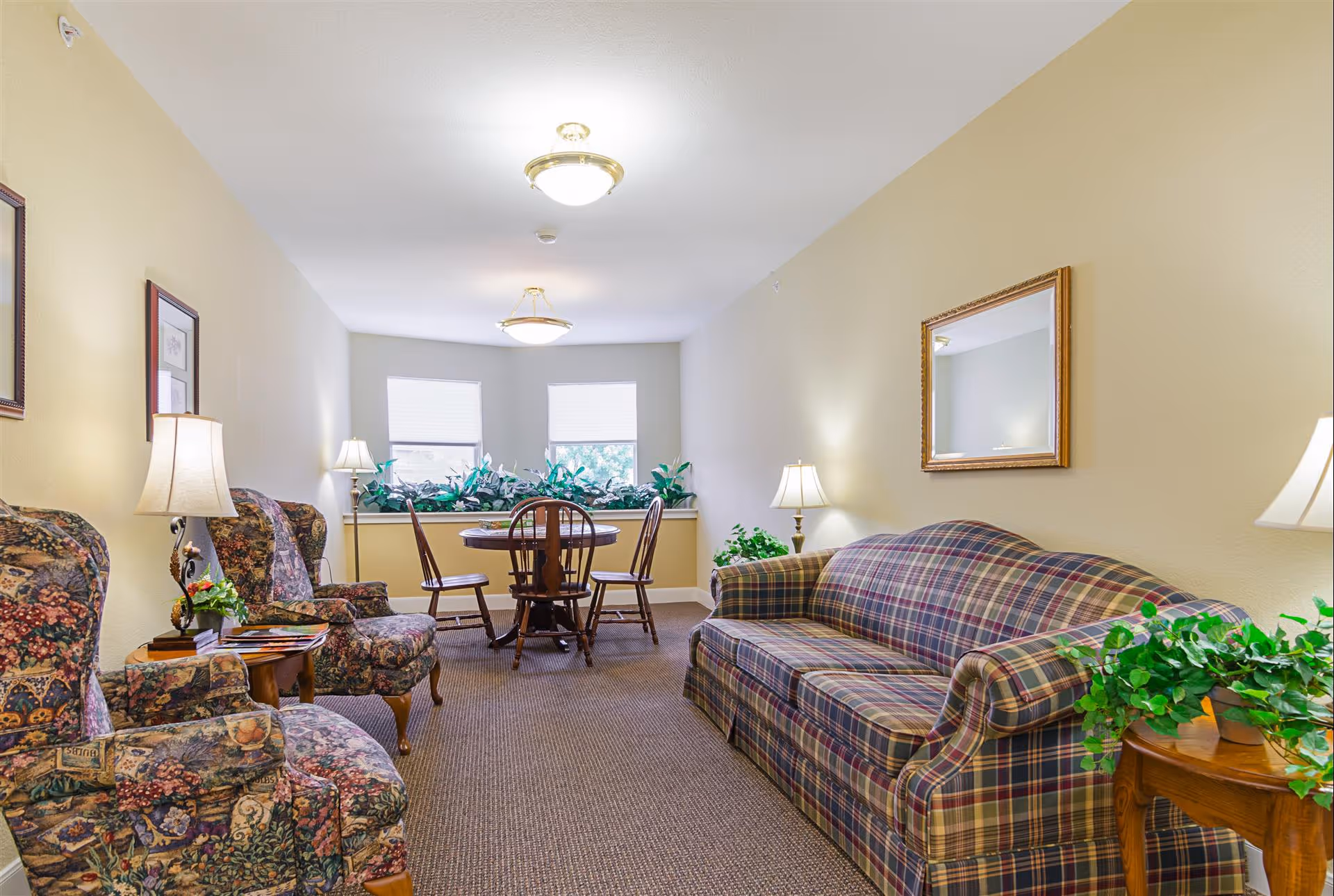 A cozy living room area in an assisted living facility with a plaid sofa, two floral upholstered armchairs, wooden side tables with lamps and plants, a round wooden dining table with four chairs near two windows, and a mirror on the wall.