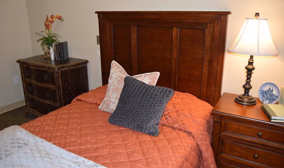 A furnished bedroom with a wooden headboard, coral quilted bedspread and decorative pillows, a bedside lamp on a nightstand and a dresser.
