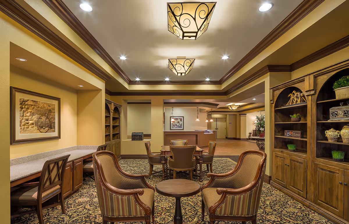 A cozy interior common area with a seating arrangement including four upholstered chairs around a small round table in the foreground and a wooden table with four chairs in the background. The room features warm yellow walls, decorative ceiling lights, built-in wooden shelves with decorative items, and a framed picture of a bicycle on the wall.
