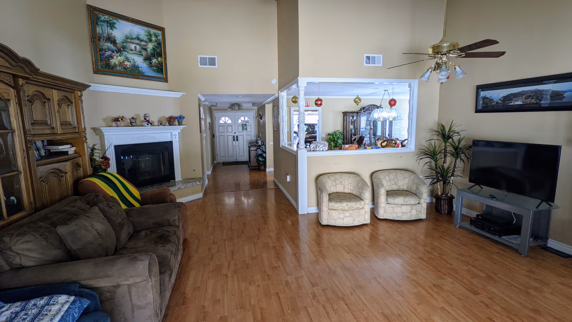 A spacious living room with wooden flooring, featuring a brown sofa, two beige armchairs, a flat-screen TV on a stand, a ceiling fan with lights, a fireplace with a white mantel, and decorative paintings on the walls. There is a large window opening into a dining area with hanging ornaments and a chandelier.
