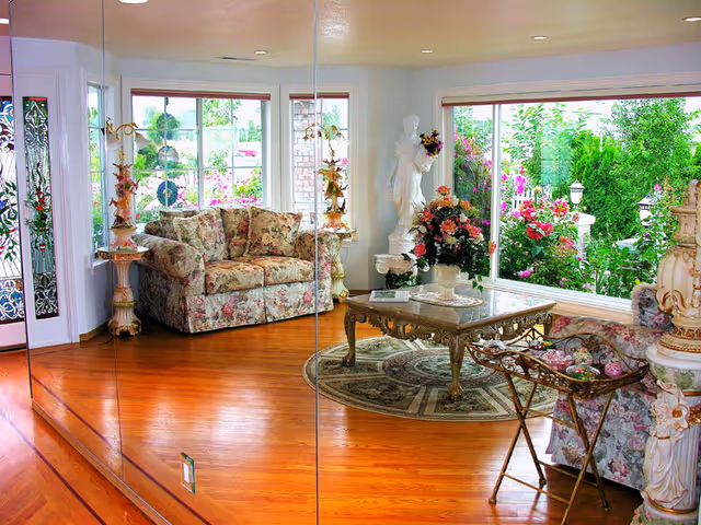 A bright living room with polished wooden floors, floral patterned sofas, a glass coffee table on a round rug, and large windows showing a colorful garden outside. The room features decorative statues and floral arrangements, with a mirrored wall reflecting the space.