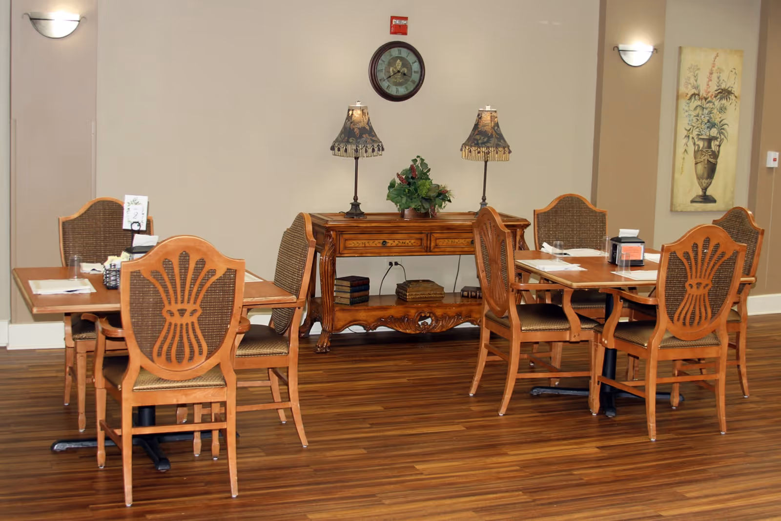 A communal dining area with wooden tables and carved chairs, a console table with lamps and a clock on the wall.