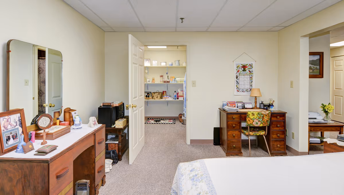 Interior view of a bedroom in Glenwood Park facility featuring a wooden dresser with a mirror on the left, a small wooden desk with a floral chair on the right, and an open door leading to a closet with shelves filled with various items. The room has beige walls and carpeted floor.