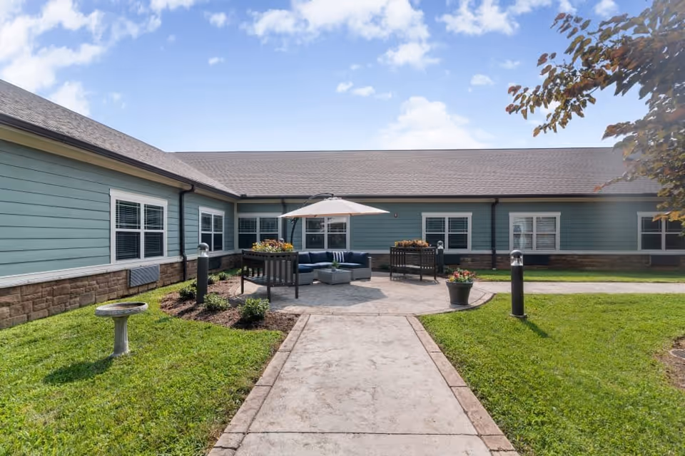 Outdoor courtyard area at Emory Ridge with a concrete pathway leading to a circular patio. The patio has a seating arrangement with a blue cushioned sectional sofa and a large umbrella. The building surrounding the courtyard has light blue siding with white-framed windows. There is green grass, a birdbath, flower pots, and some small plants around the patio under a partly cloudy sky.