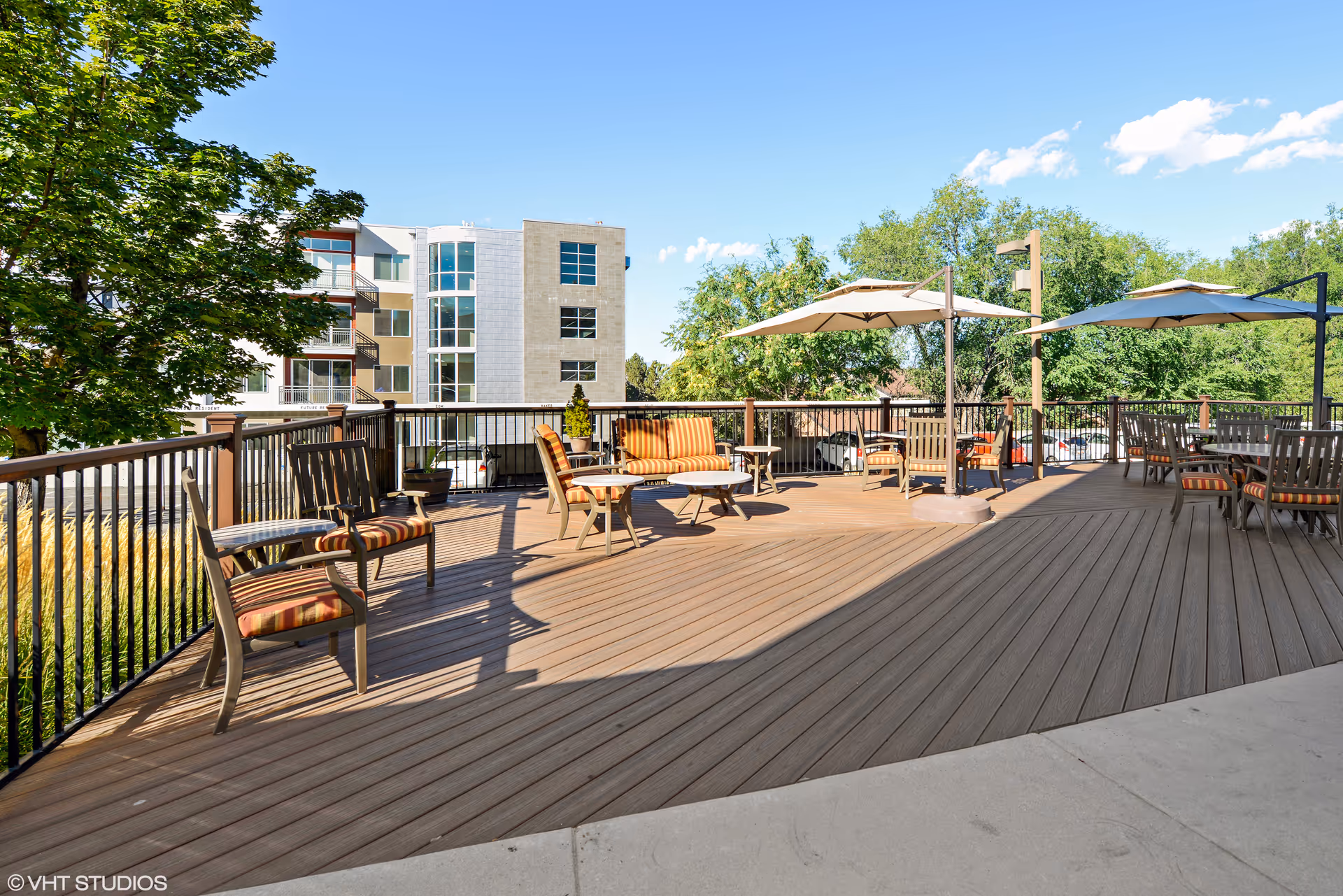 Sunlit outdoor deck with chairs, tables, and umbrellas overlooking trees and a nearby building.