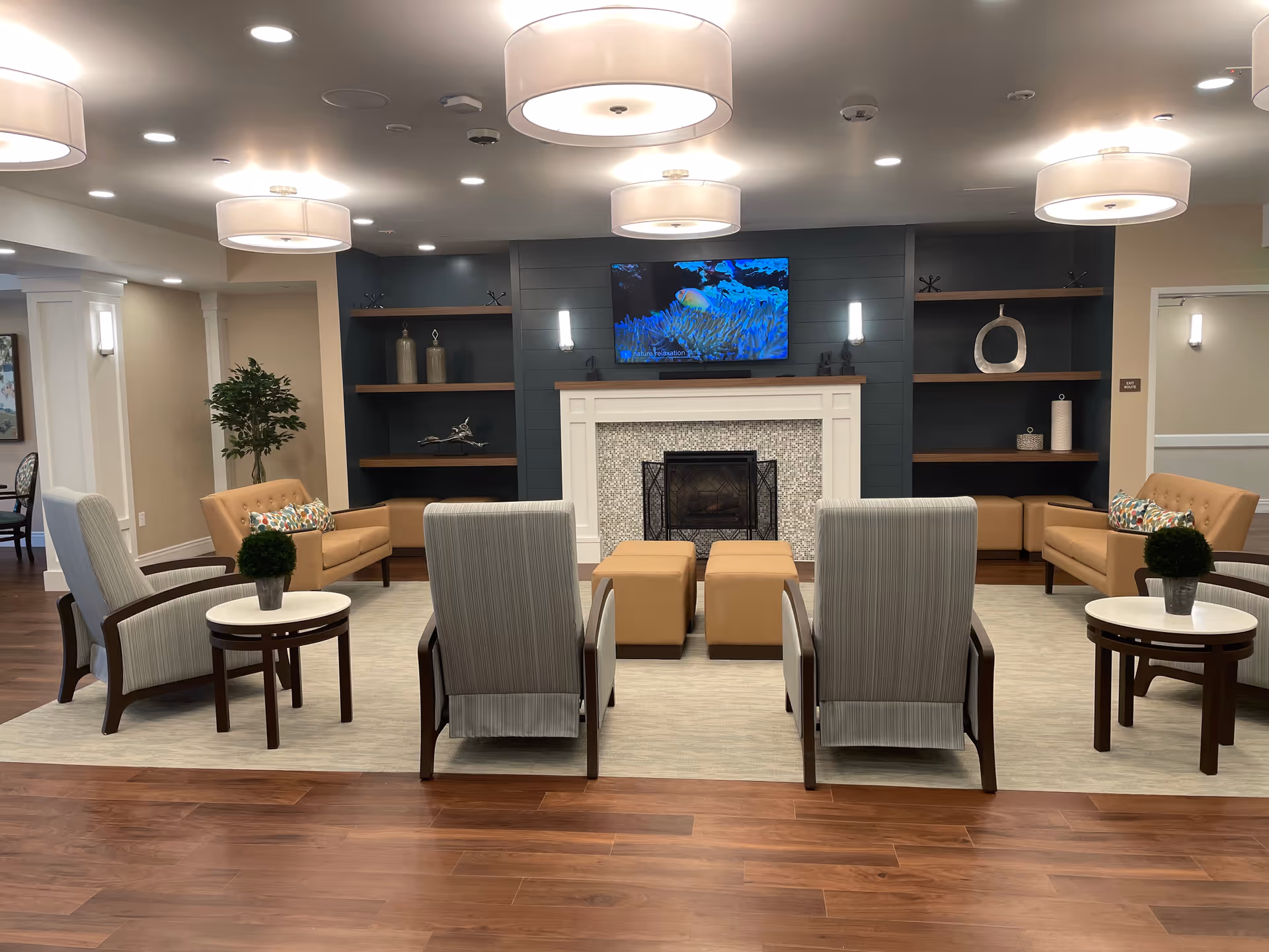 A modern living room area in an assisted living facility featuring a fireplace with a mounted flat-screen TV above it. The room has four armchairs with striped upholstery, two small round tables with potted plants, two beige sofas with colorful pillows, and two beige ottomans in front of the fireplace. The walls have built-in shelves with decorative items, and the ceiling has multiple round light fixtures. The floor is wooden with a large area rug in the seating area.