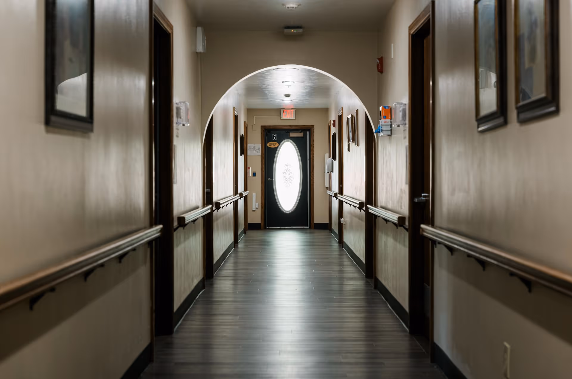 A long, empty hallway in an assisted living facility with wooden handrails on both sides, several closed doors, framed pictures on the walls, and an arched ceiling near the end leading to a door with an oval frosted glass window.