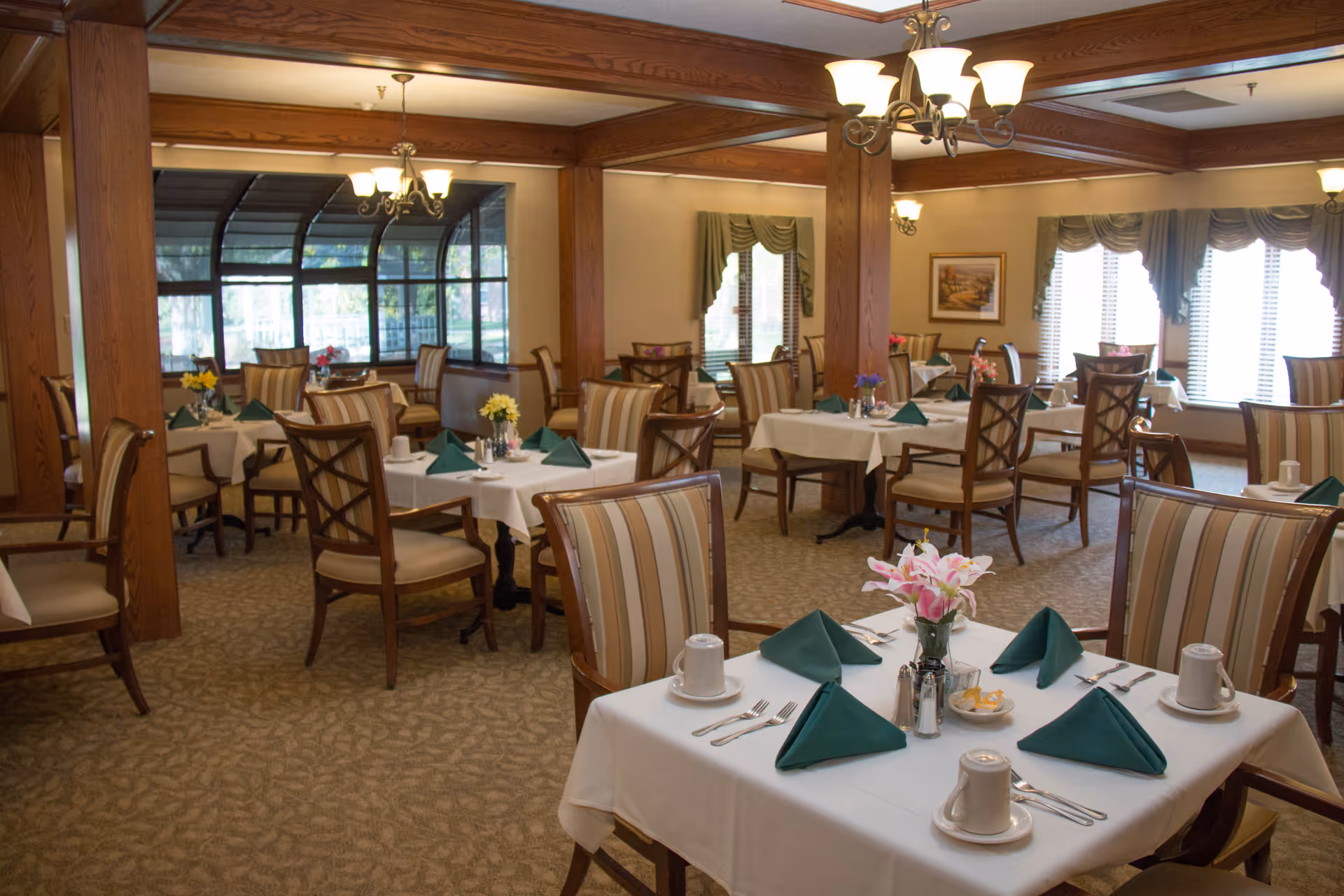 Dining room with several tables set with white linens, green napkins, floral centerpieces and wooden chairs.