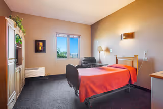 A simple, clean bedroom in a senior living facility with a single bed covered in a red blanket, a wooden wardrobe, a black armchair near a window, and beige walls with soft lighting.