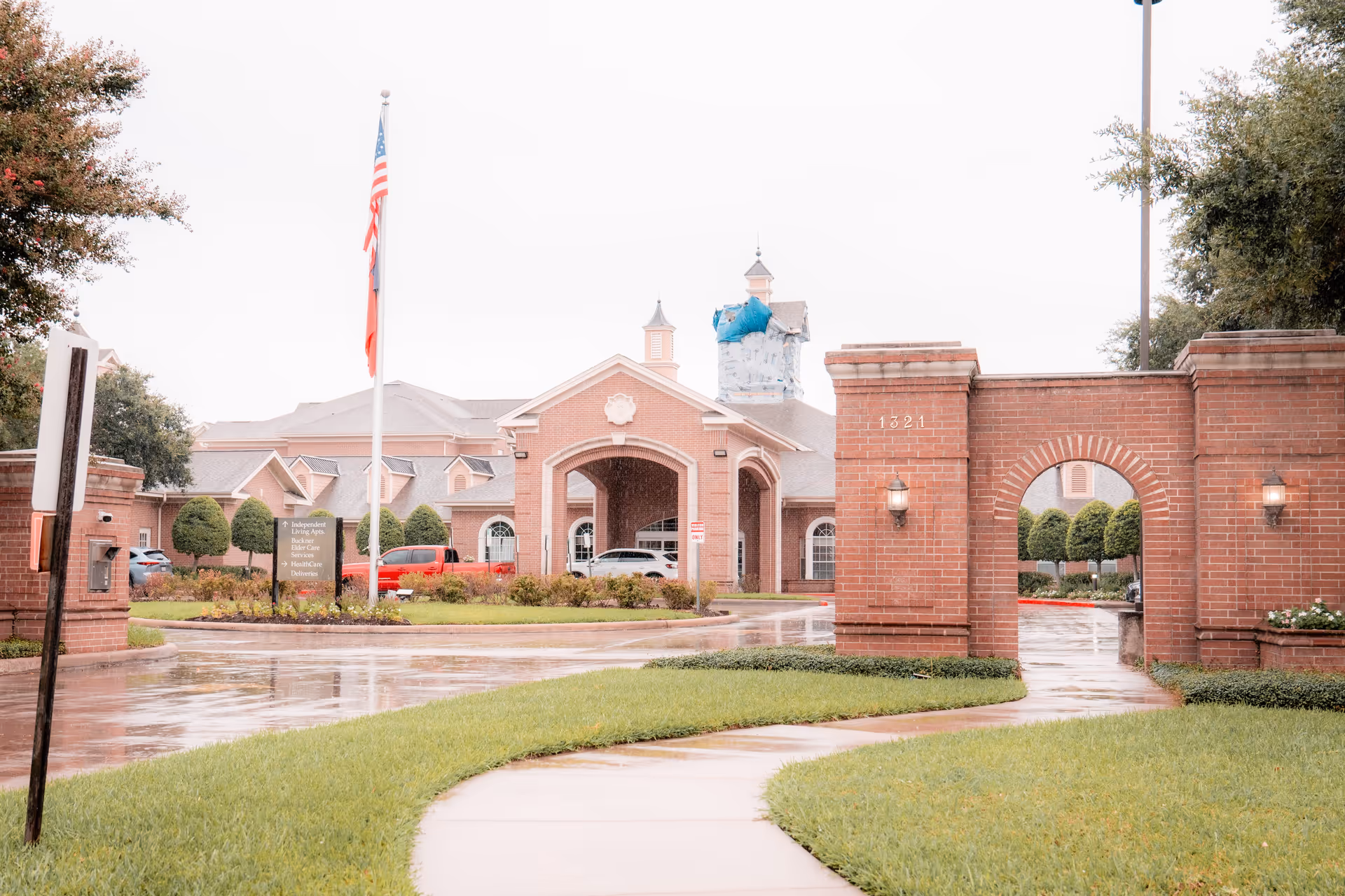 Entrance to Buckner Parkway Place featuring a brick archway with the number 1321, a curved sidewalk, green grass, an American flag on a flagpole, and a large building with a covered driveway in the background on a rainy day.