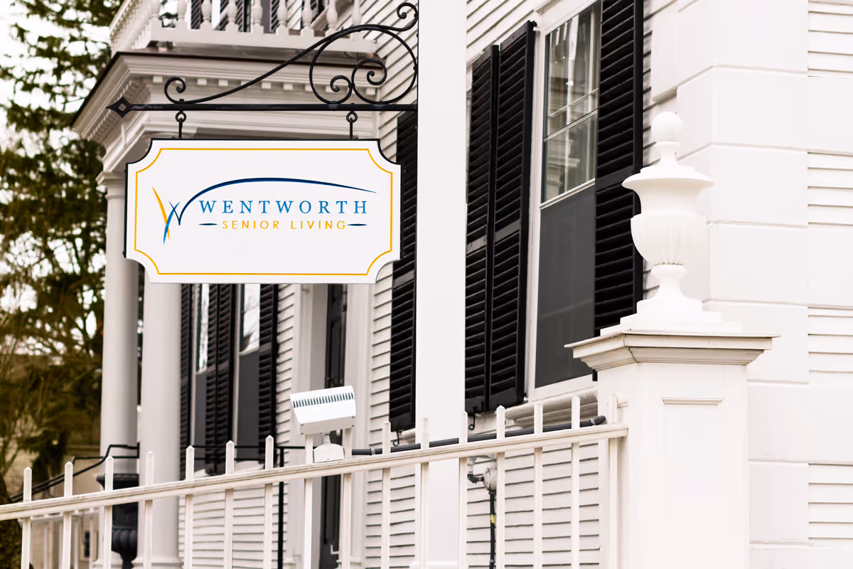 A hanging Wentworth Senior Living sign on the exterior of a white building with black shutters and a decorative railing.