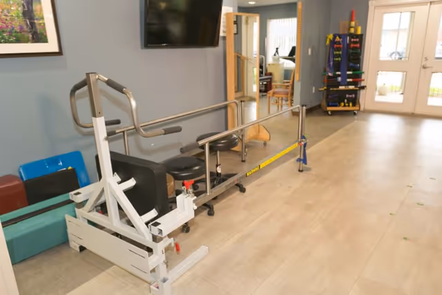 An interior room with physical therapy equipment including parallel bars for walking exercises, stools, and a rack of colorful weights. There is a large TV mounted on the wall and double glass doors leading outside.