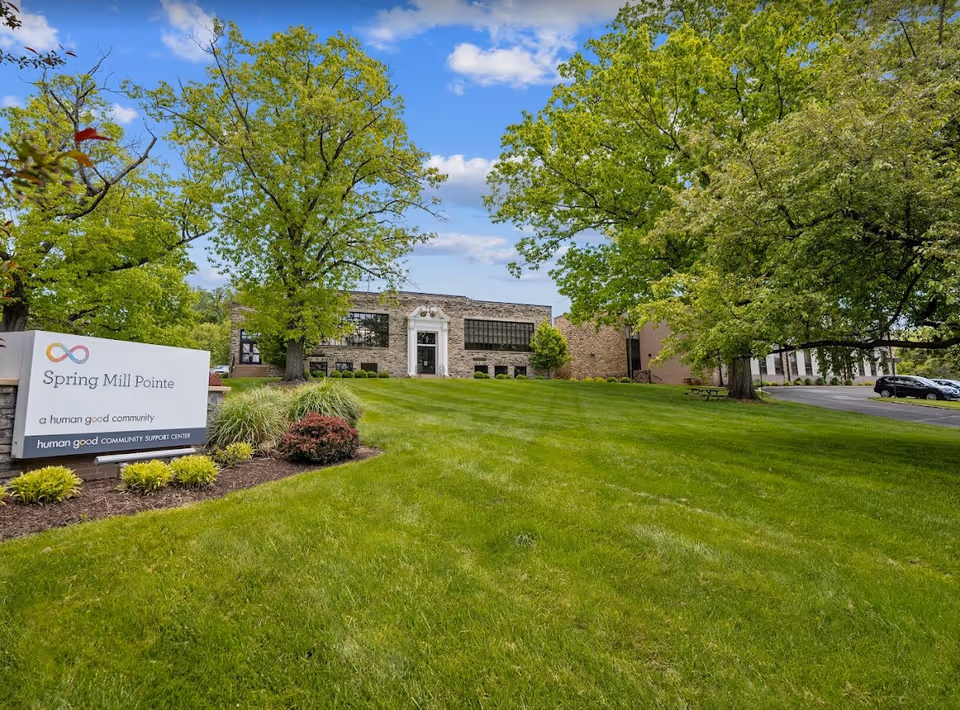 Exterior view of Spring Mill Pointe facility with a large green lawn, trees, and a stone building in the background under a partly cloudy blue sky. A sign in the foreground reads 'Spring Mill Pointe, a human good community, human good COMMUNITY SUPPORT CENTER.'