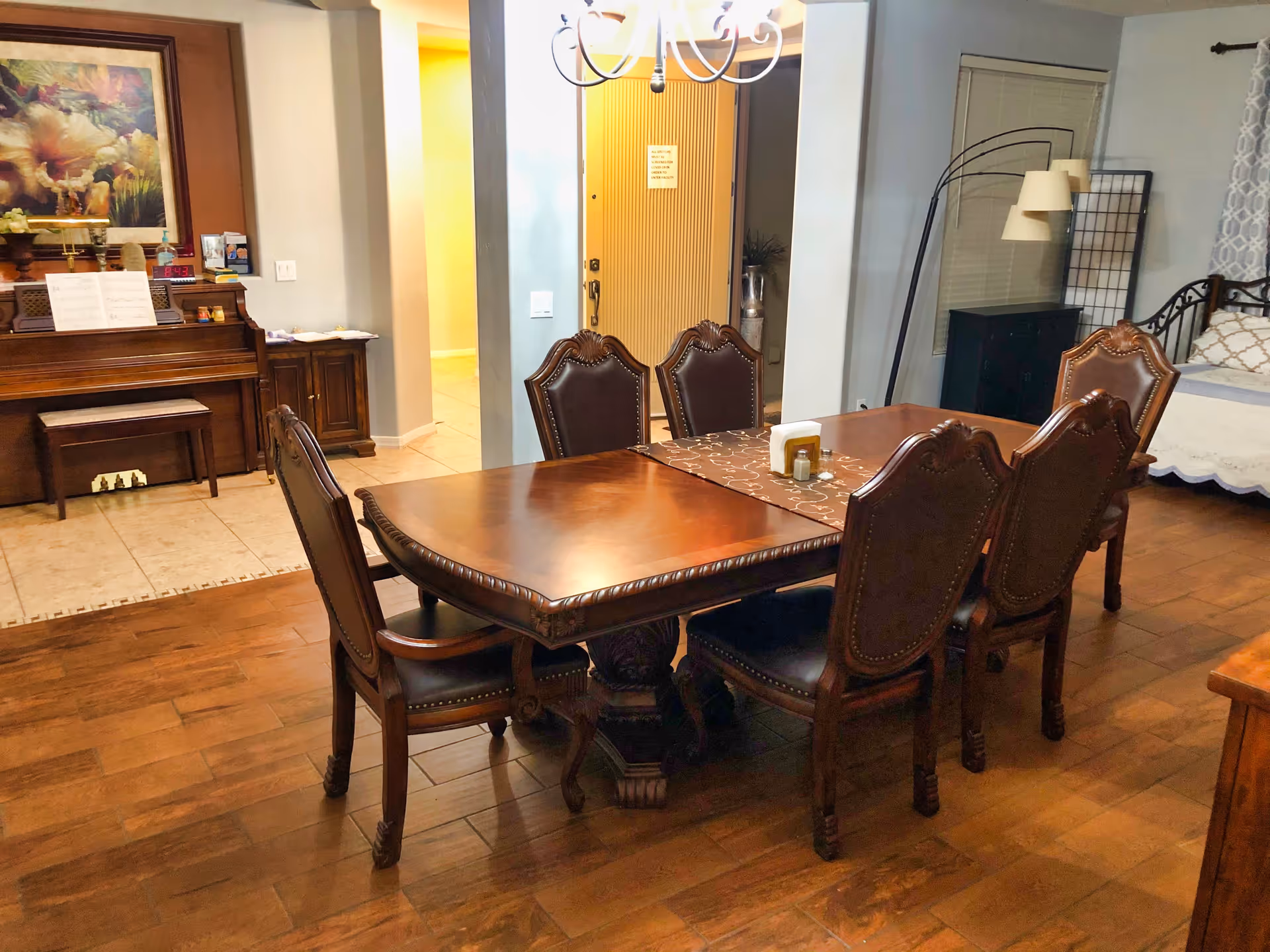 A dining area with a wooden table and six ornate chairs. In the background, there is a piano with sheet music, a painting above it, and a small cabinet. To the right, there is a bed with pillows, a floor lamp, and a window with blinds and curtains. The floor is wooden, and there is a chandelier hanging above the table.