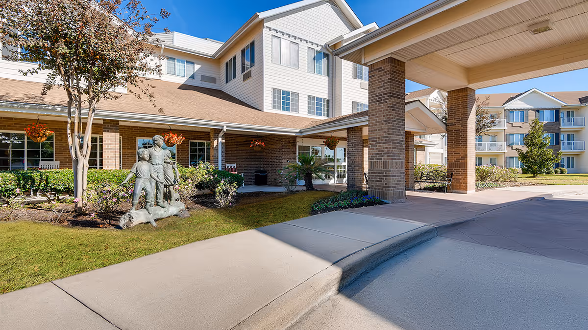 Exterior view of Copperfield Estates - Sky Active Living showing a covered entrance with brick pillars, manicured lawn, a statue of three children and a dog, and multi-story residential buildings in the background under a clear blue sky.