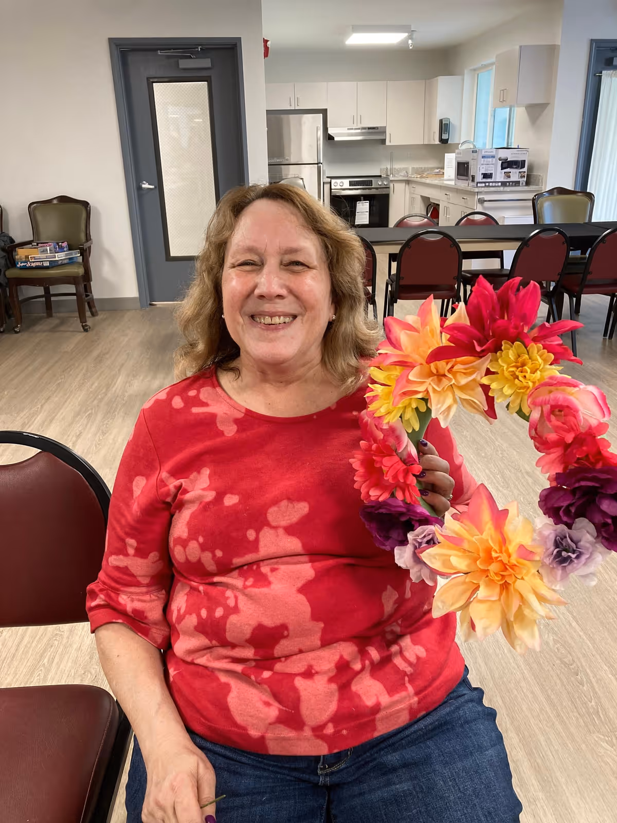 A smiling woman wearing a red patterned shirt is seated indoors, holding a colorful floral wreath. Behind her is a kitchen area with white cabinets, a refrigerator, stove, microwave, and a dining table with chairs.