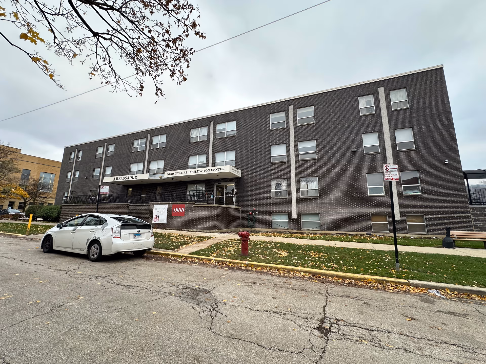 Three-story dark brick building labeled "Ambassador Nursing & Rehabilitation Center" facing the street with a parked white car, sidewalk, and lawn.