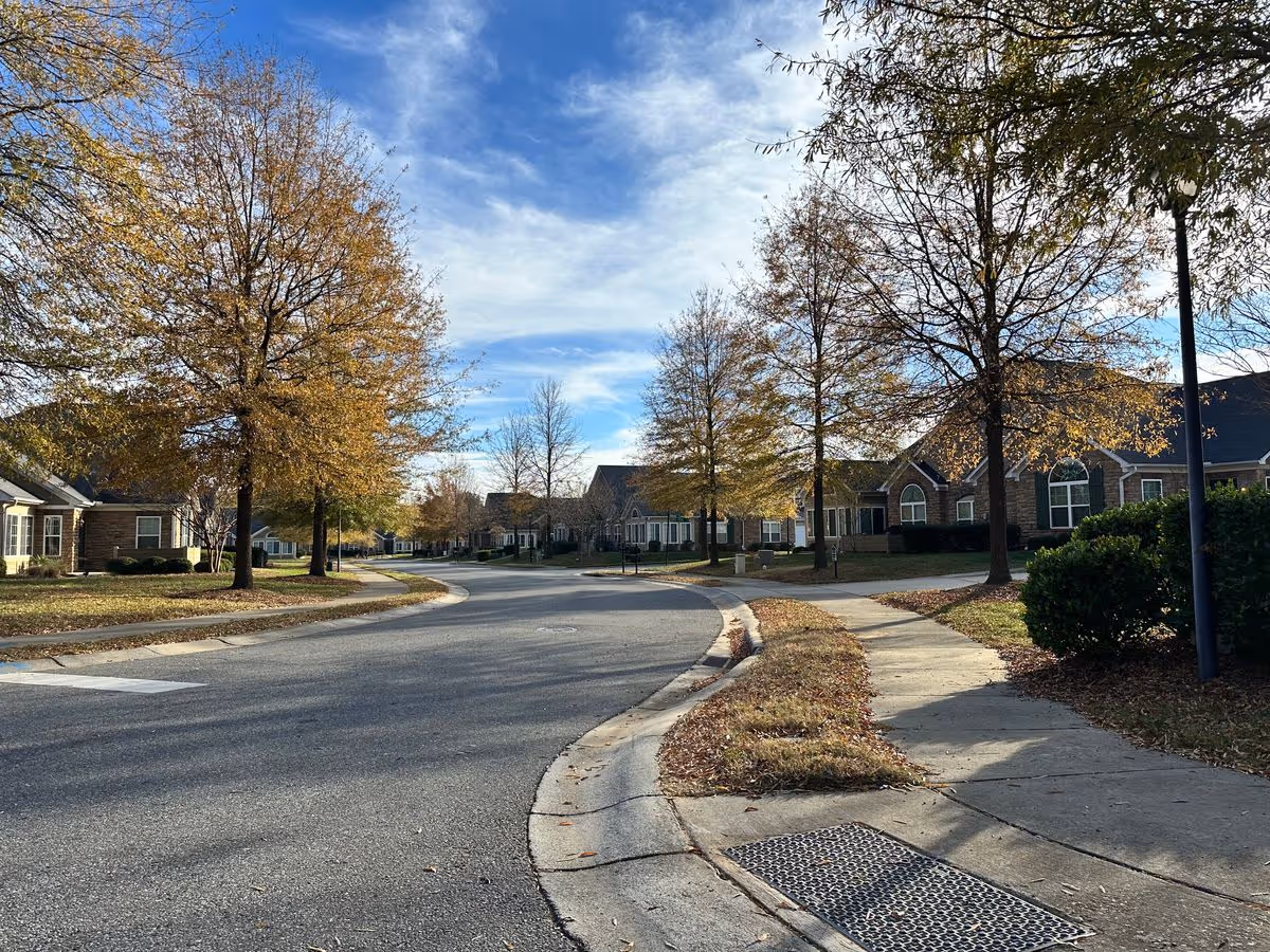 Curving residential street lined with single-story brick villas and trees with autumn foliage under a blue sky.