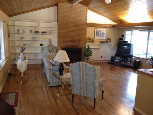 Spacious living room with a vaulted wood ceiling, brick fireplace, built-in shelving, sofa and armchair, and a TV by a large window.