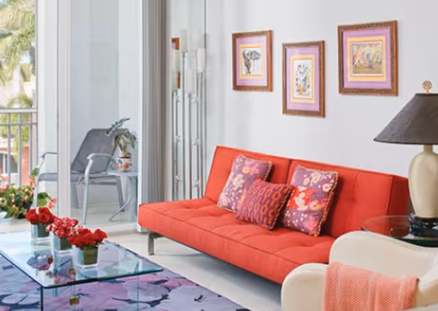 A bright living room featuring a red sofa with three decorative pillows, a glass coffee table with red flowers in vases, a white armchair with a pink throw blanket, a table lamp with a dark shade, and three framed artworks on the wall. Sliding glass doors open to a balcony with outdoor chairs and a small table.