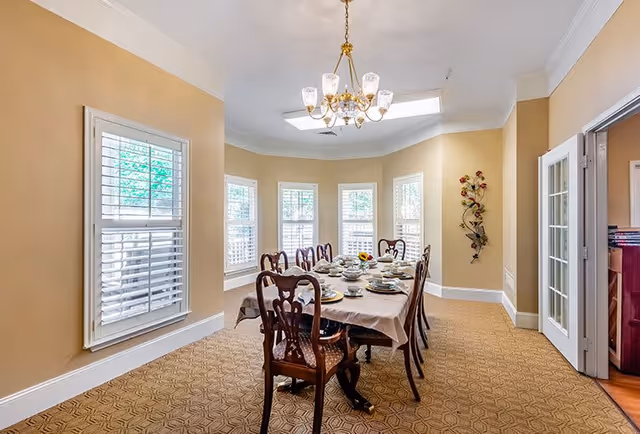 A well-lit dining room with a rectangular table covered with a beige tablecloth and set with plates, cups, and utensils. The room has beige walls, a patterned carpet, multiple windows with white shutters, and a chandelier hanging from the ceiling. There is a decorative wall piece with flowers on one wall and double glass doors leading to another room.