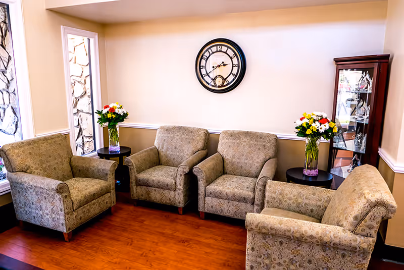A cozy seating area with four patterned armchairs arranged around side tables holding vases of flowers, a wall clock, and a glass display cabinet.