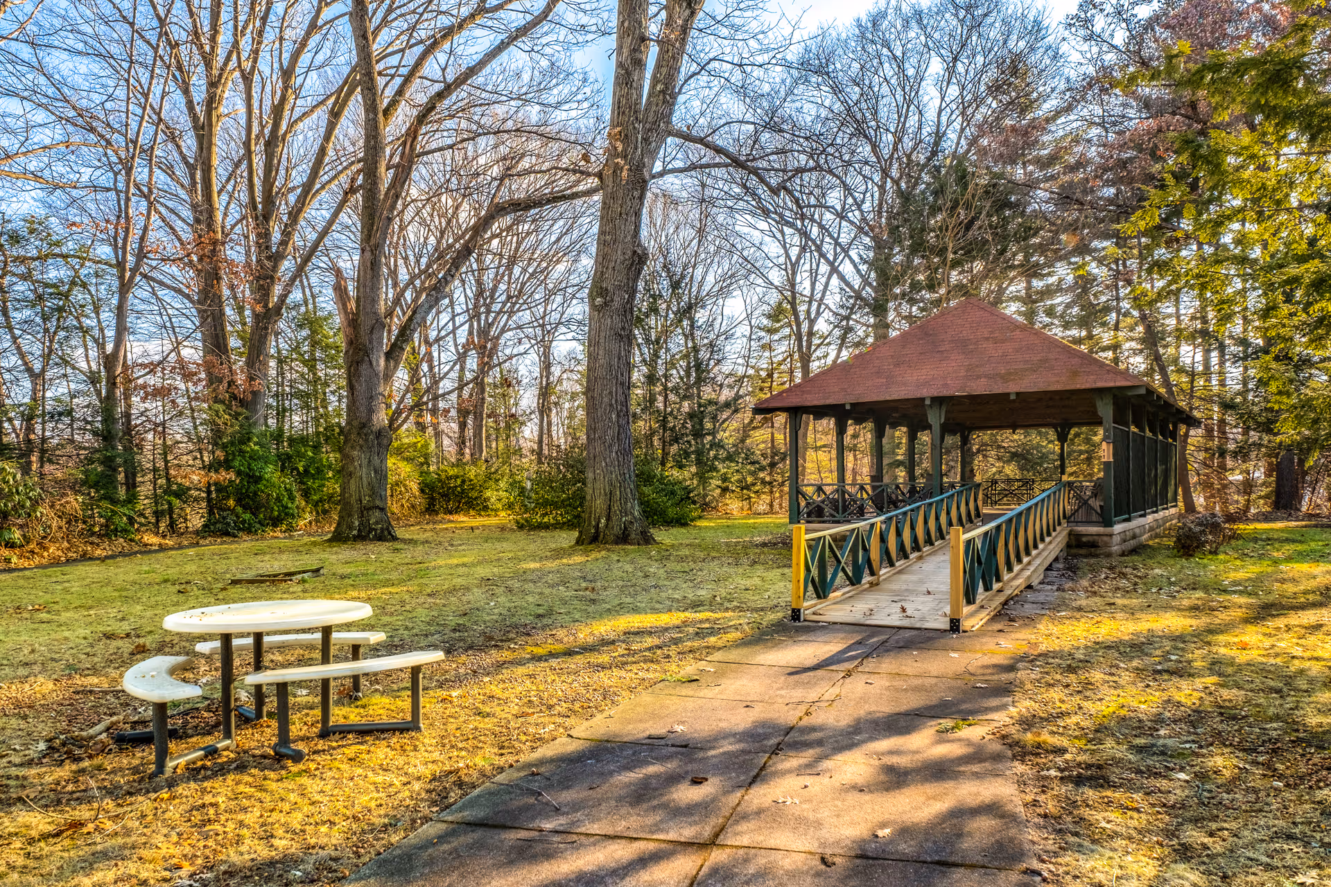 Outdoor scene at a senior living facility featuring a paved walkway leading to a covered wooden gazebo with a red roof. The area is surrounded by tall trees with bare branches and some green shrubs. A round picnic table with attached benches is situated on the grass to the left of the walkway.