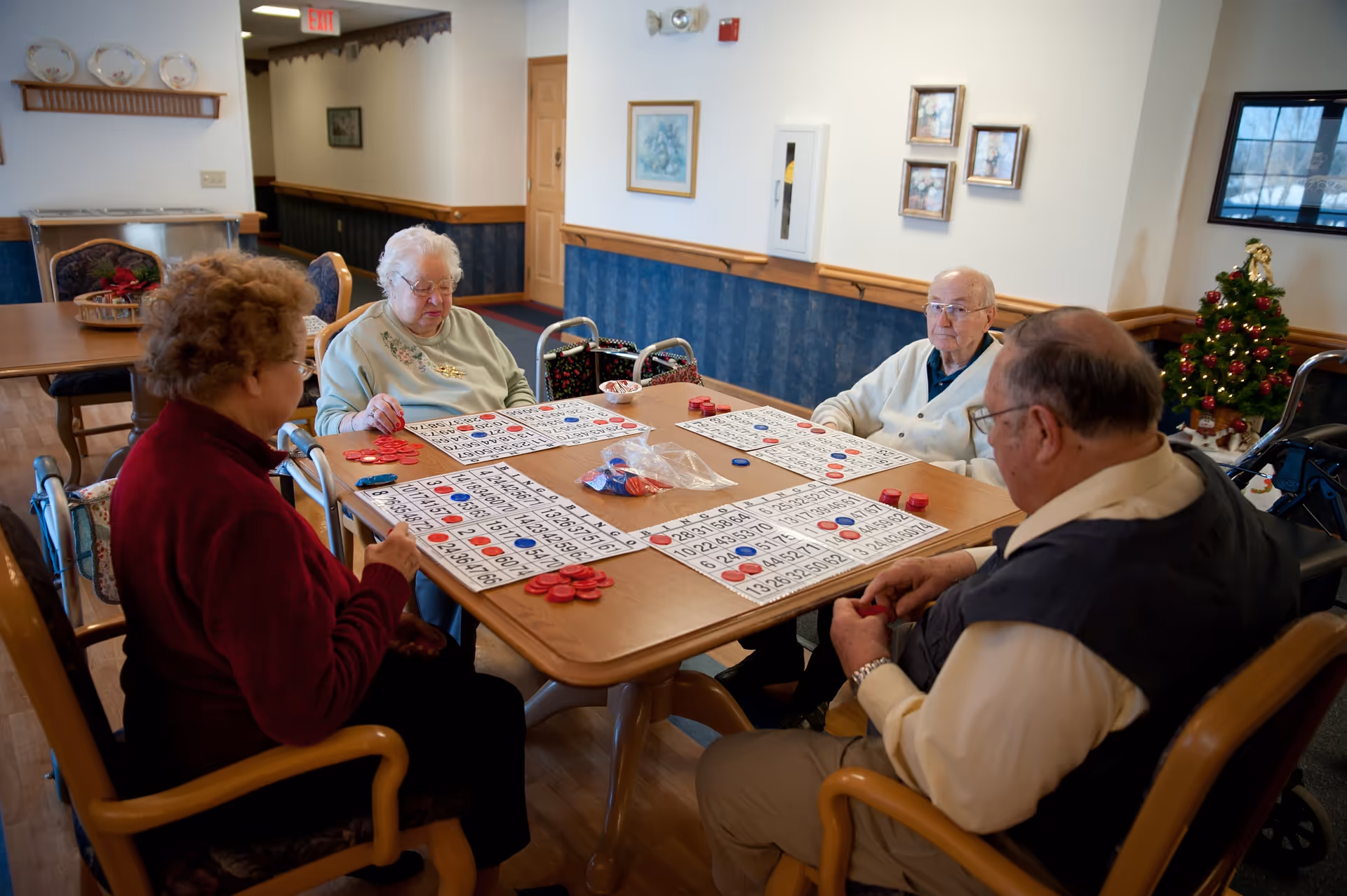 Four elderly people sitting around a wooden table playing bingo in a well-lit room with blue and white walls. There is a small decorated Christmas tree in the corner and framed pictures on the wall.