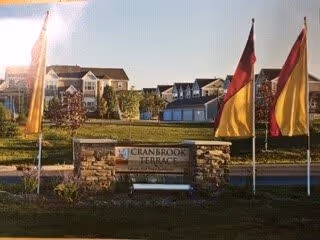 Stone entrance sign reading "Cranbrook Terrace" with flagpoles, a bench, lawn, and the facility buildings in the background.