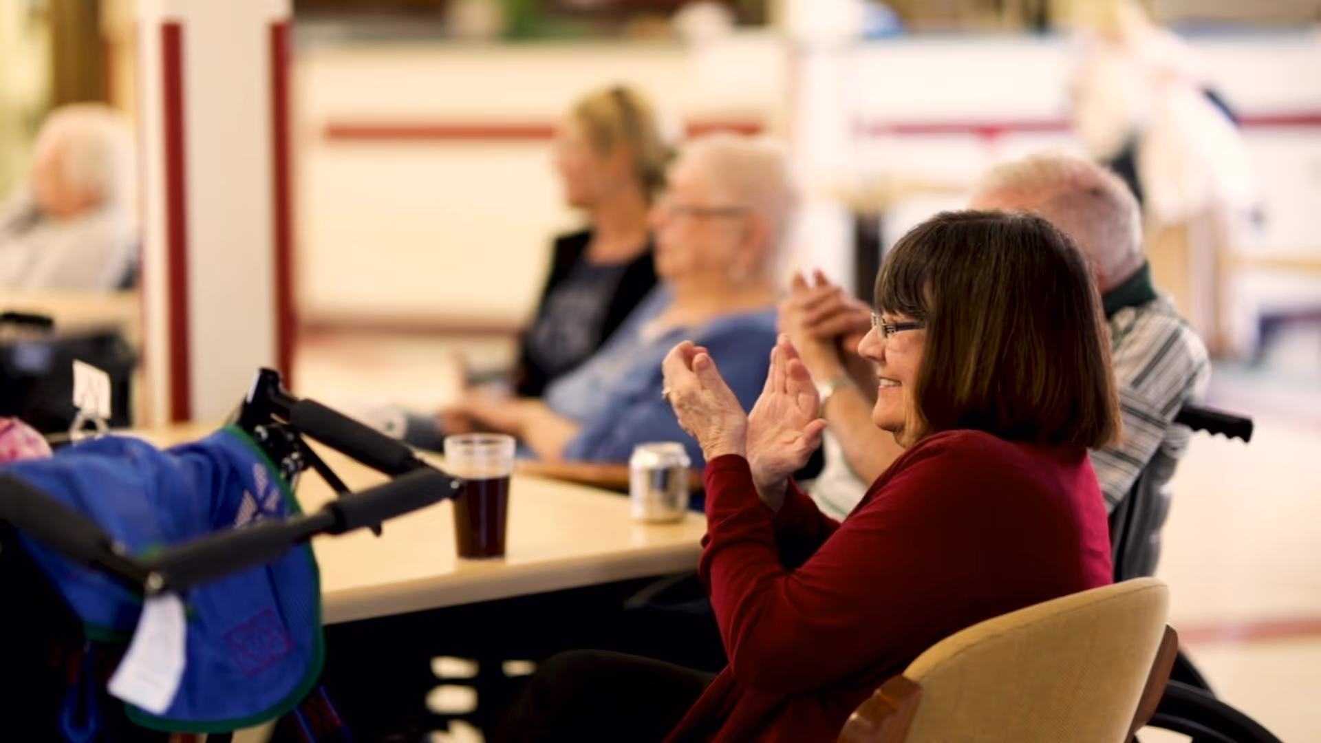 A group of elderly people sitting around a table in a common area, with one woman in the foreground clapping and smiling. There are drinks on the table and a walker nearby.