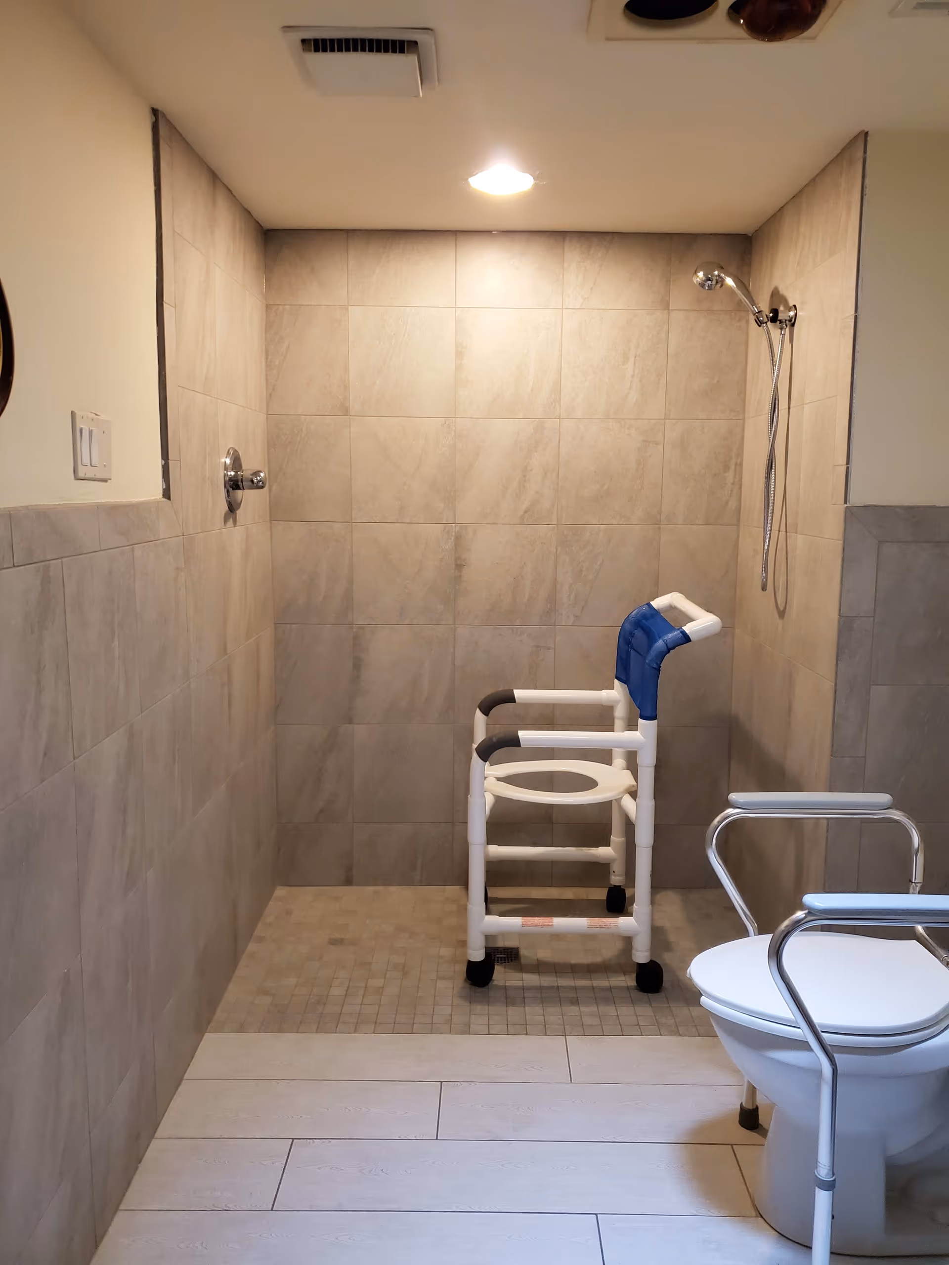 Tiled accessible bathroom featuring a walk-in shower with a shower chair and a raised toilet frame.