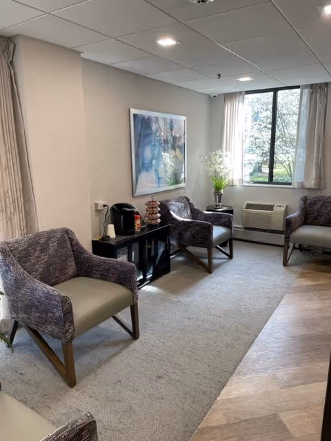A cozy sitting area in a senior living facility with three upholstered armchairs arranged around a small black cabinet. The cabinet holds a coffee maker and a decorative item. A large window with light curtains lets in natural light, and a framed abstract painting hangs on the wall above the cabinet. The floor is a combination of carpet and wood-like flooring.