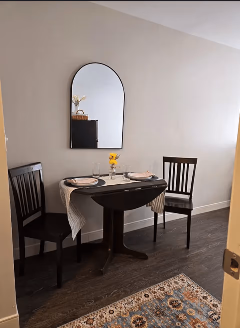 Small dining area with a round drop-leaf table set for two, two wooden chairs, a wall mirror, and a patterned rug.
