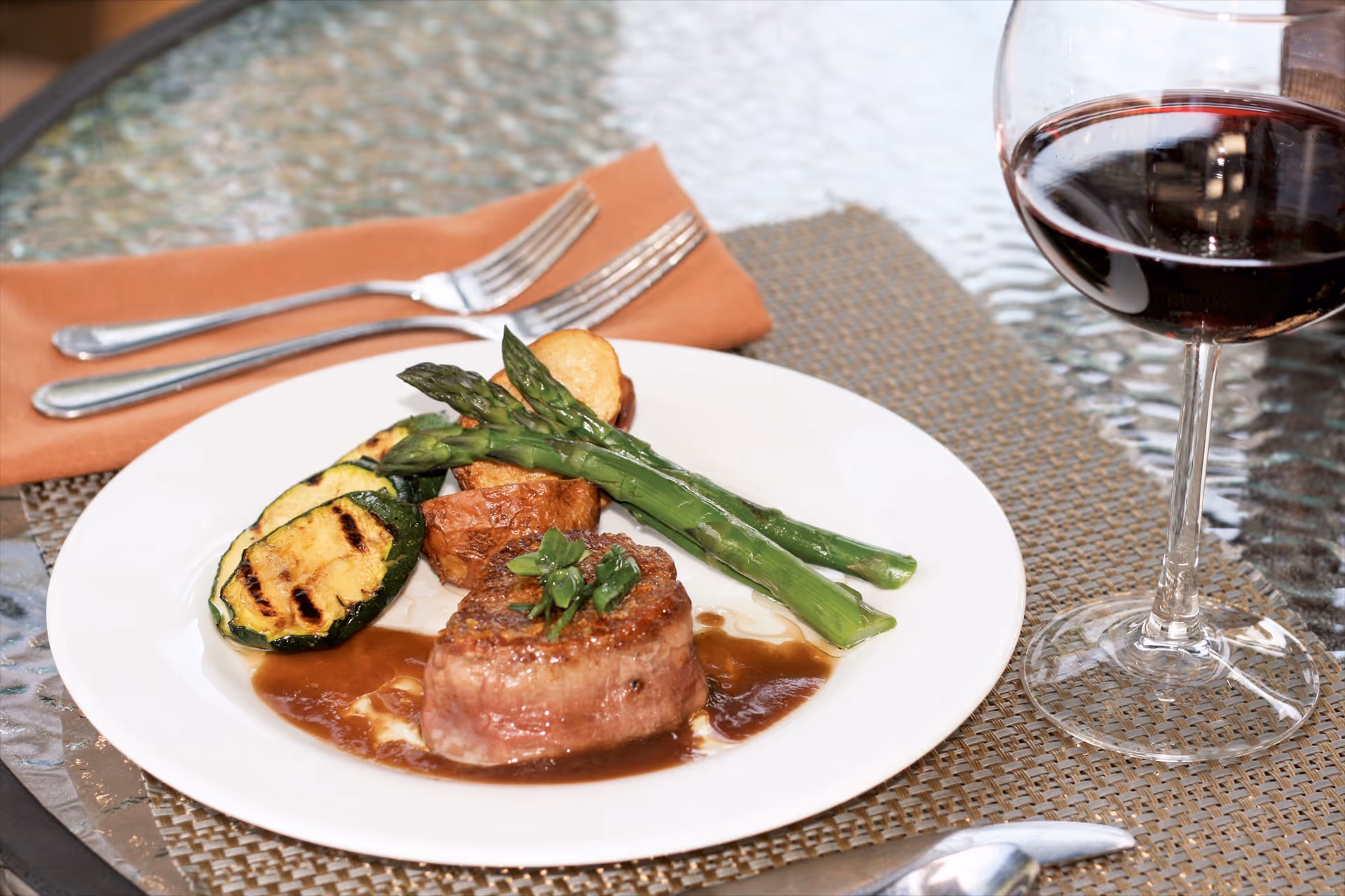 A plated steak with asparagus, grilled zucchini and potatoes beside a glass of red wine on a woven placemat.