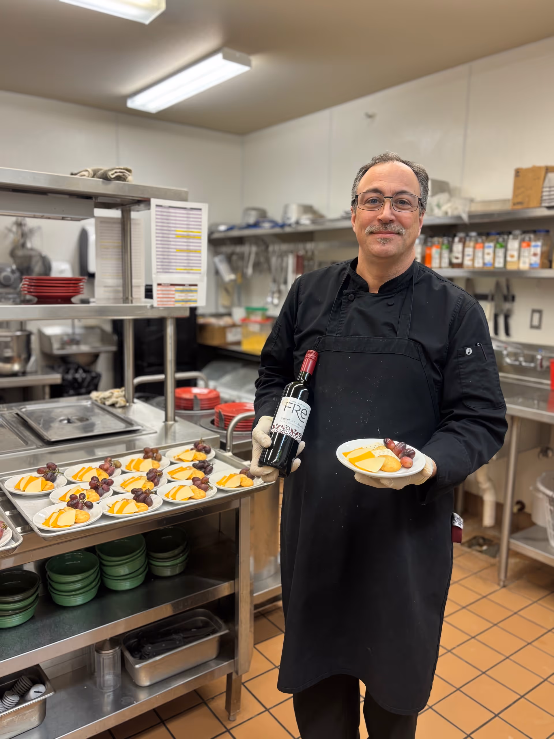 A man wearing a black chef's coat and apron stands in a commercial kitchen holding a plate with cheese, grapes, and crackers in one hand and a bottle of red wine in the other. Behind him, there is a metal rack with multiple plates of similar cheese and grape arrangements. The kitchen has stainless steel counters, shelves with spices, and various kitchen utensils.