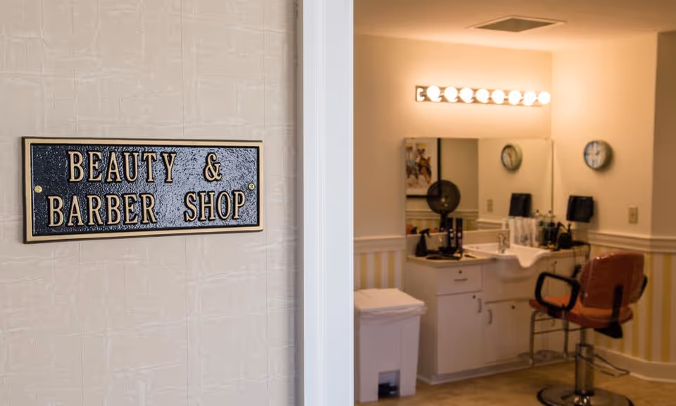 Interior view of a beauty and barber shop in a senior living facility, showing a salon chair, a large mirror with lights above it, a sink, and various hair styling tools on the counter.