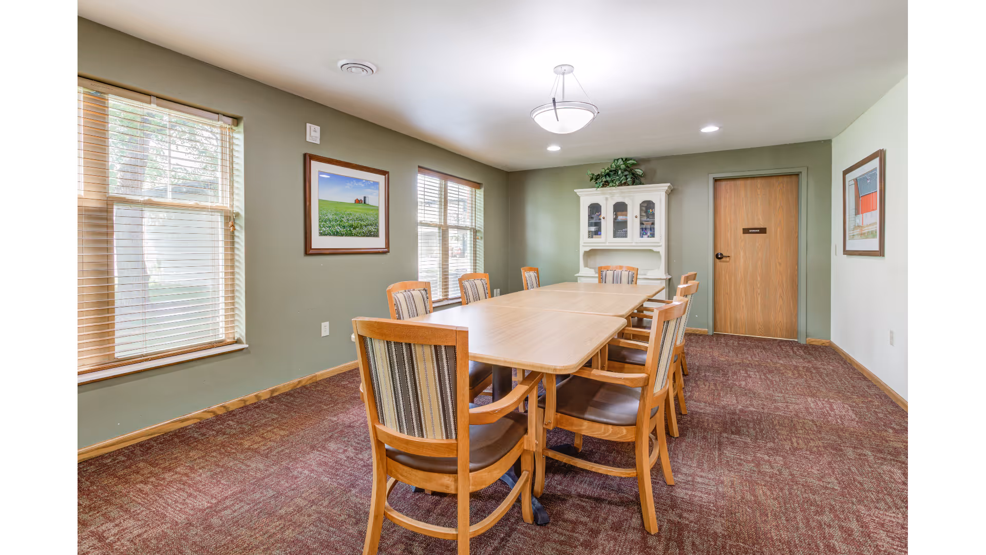 Small dining/meeting room with a long wooden table surrounded by chairs, windows on the left, a white hutch and a closed door at the back.