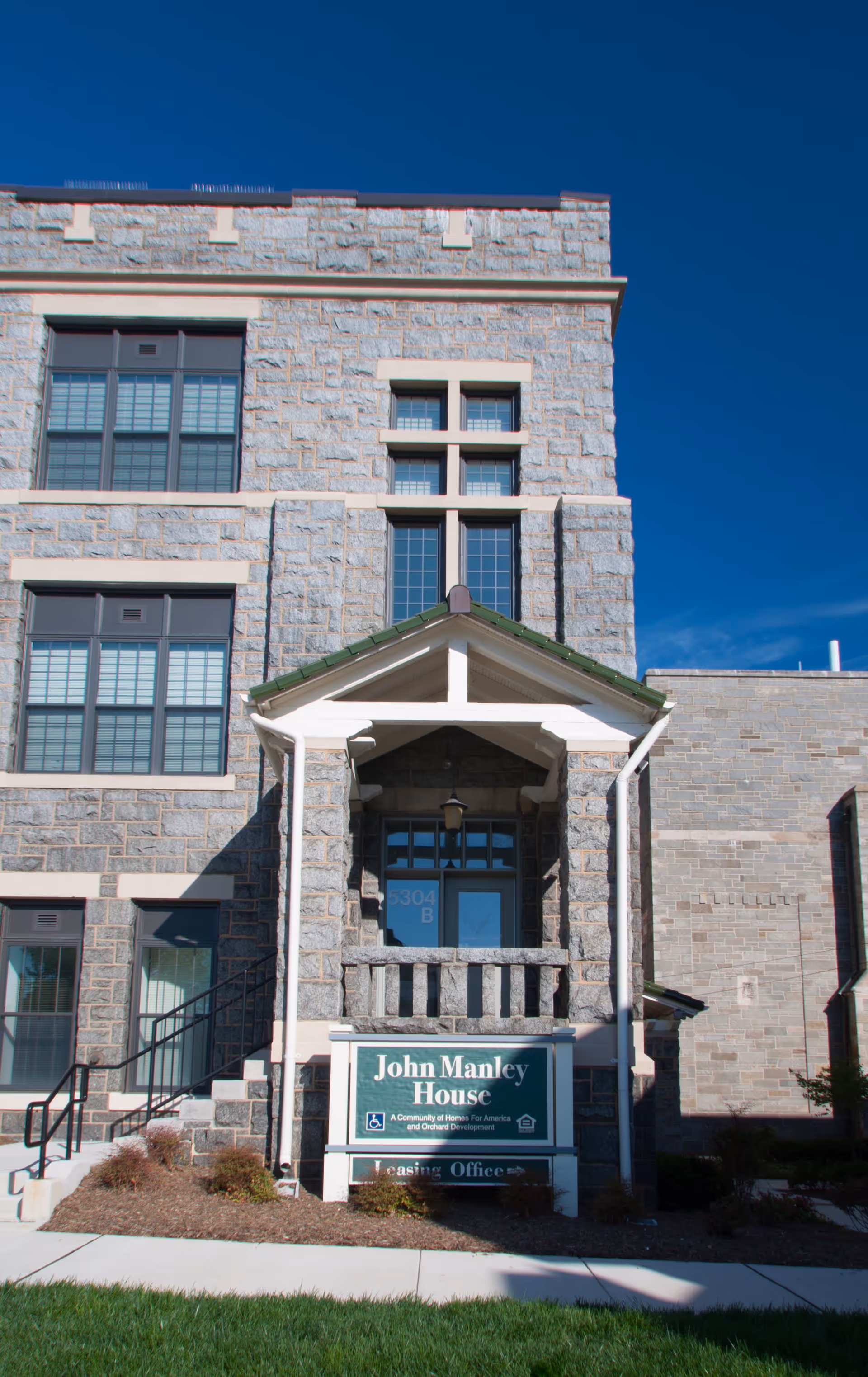 Exterior front view of John Manley House, a stone building with large windows and a small covered porch above the entrance. A green sign in front reads 'John Manley House, A Community of Homes For America and Orchard Development, Leasing Office'.