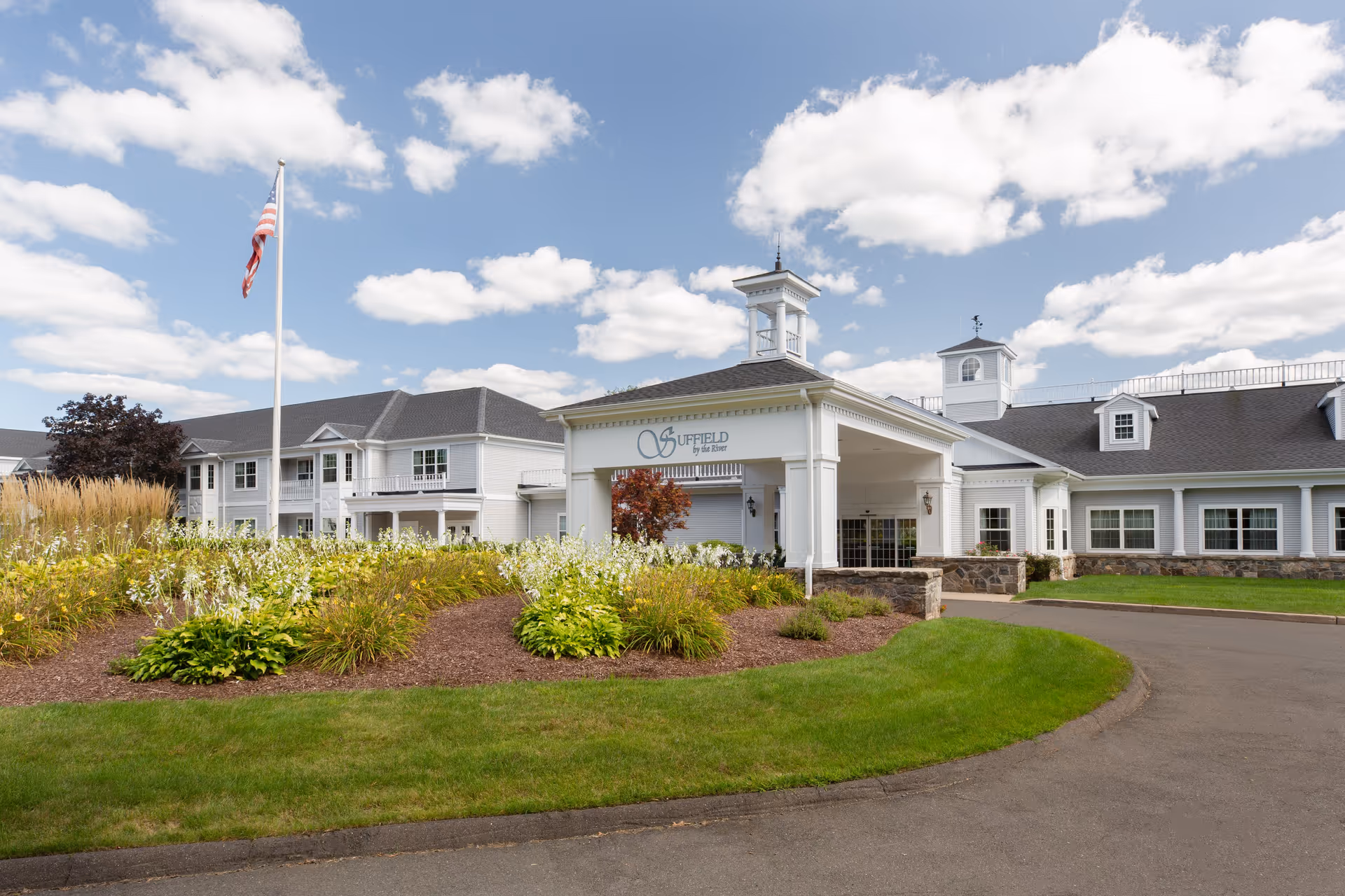 Exterior of Suffield by the River senior living facility with landscaped entrance and flag.
