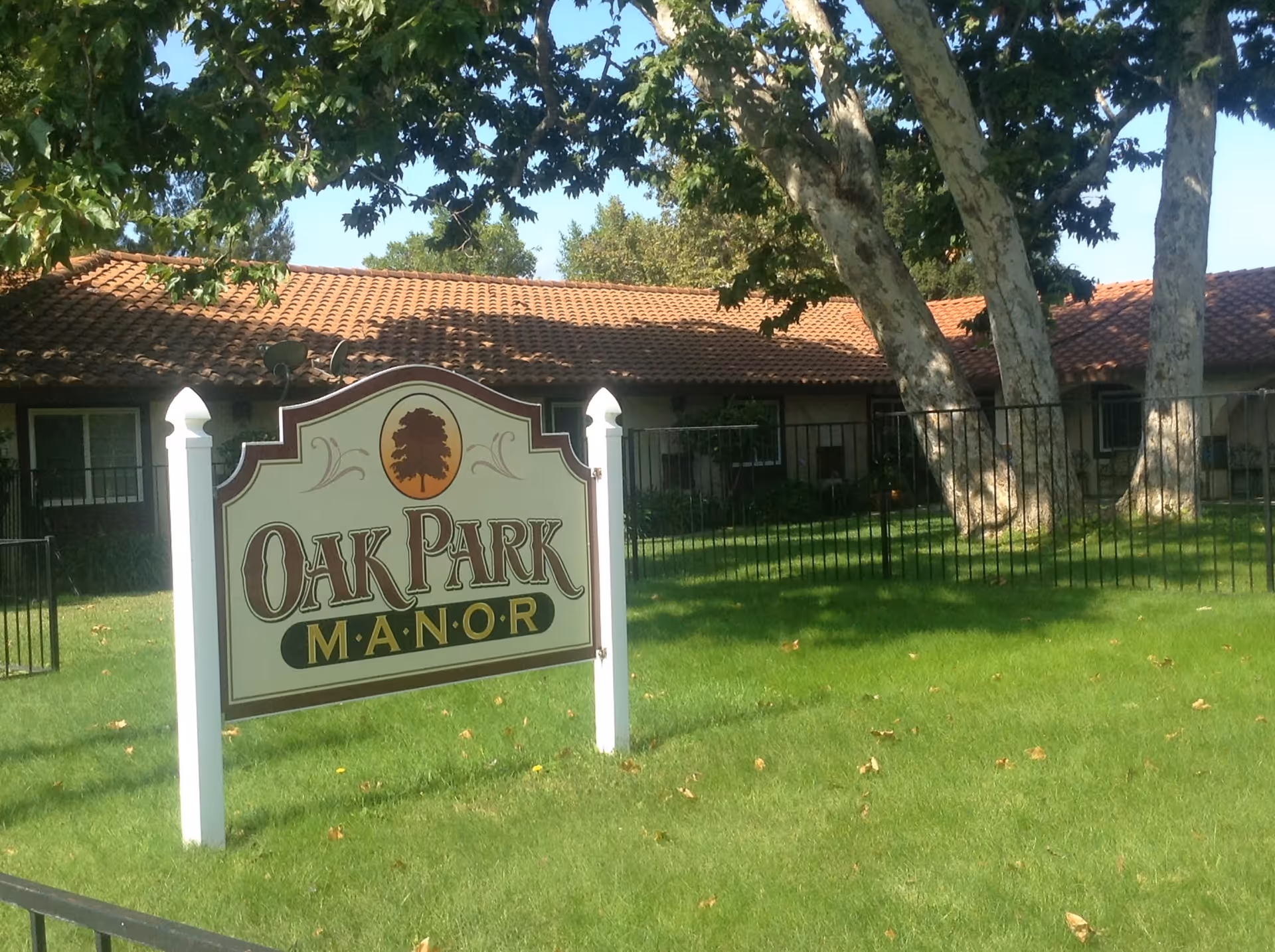 A white sign that reads "Oak Park Manor" on a green lawn in front of a single-story building with a tiled roof and large trees.