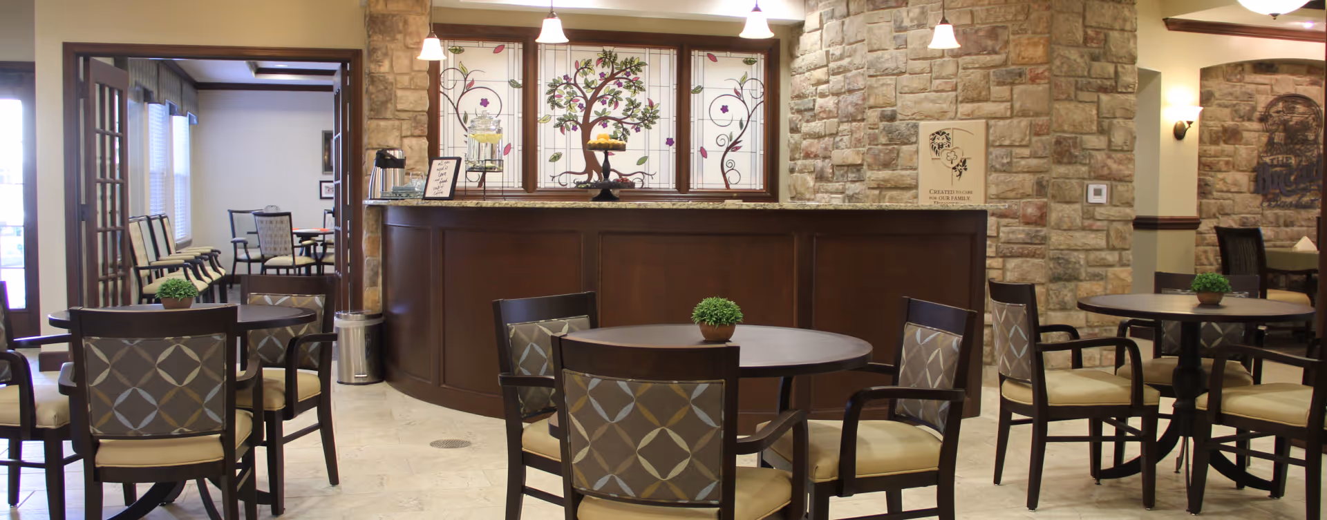 A cozy dining area in a senior living facility featuring round tables with patterned cushioned chairs. The room has a stone accent wall and a wooden counter with decorative stained glass panels behind it. Small potted plants are placed on each table, and pendant lights hang from the ceiling providing warm lighting.