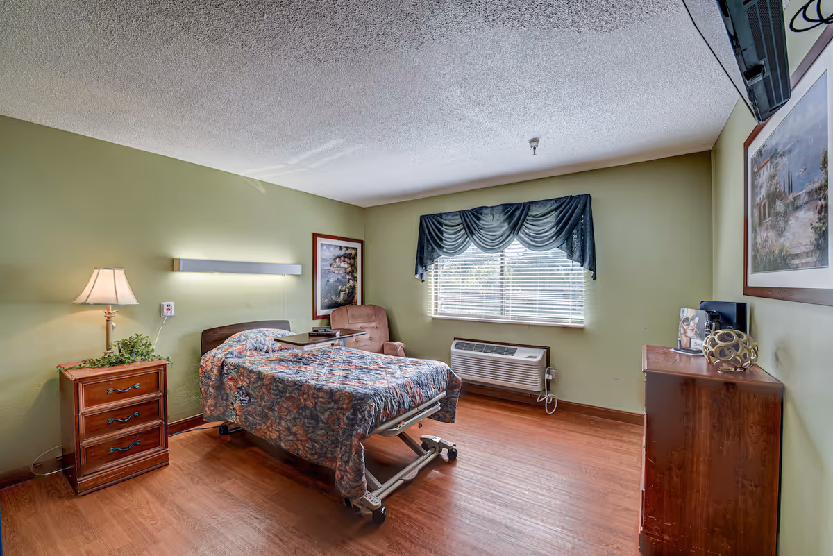 A senior living facility bedroom with a hospital-style bed covered in a floral bedspread, a wooden nightstand with a lamp and plant, a comfortable armchair, a window with blinds and a dark valance, a wall-mounted air conditioning unit, a wooden dresser with decorative items, and framed artwork on the walls. The room has light green walls and wood flooring.