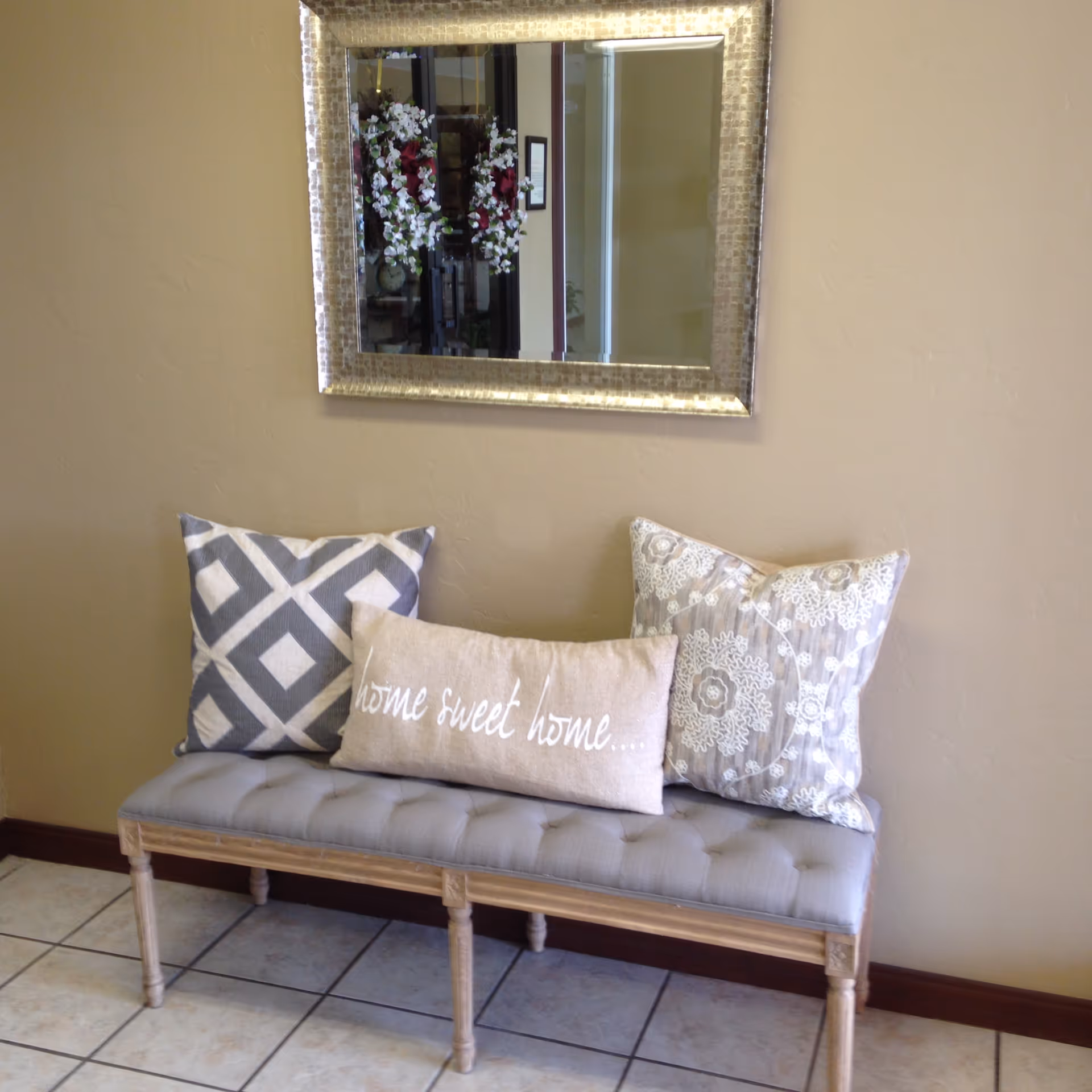 Tufted bench with three decorative pillows beneath a framed mirror on a beige wall