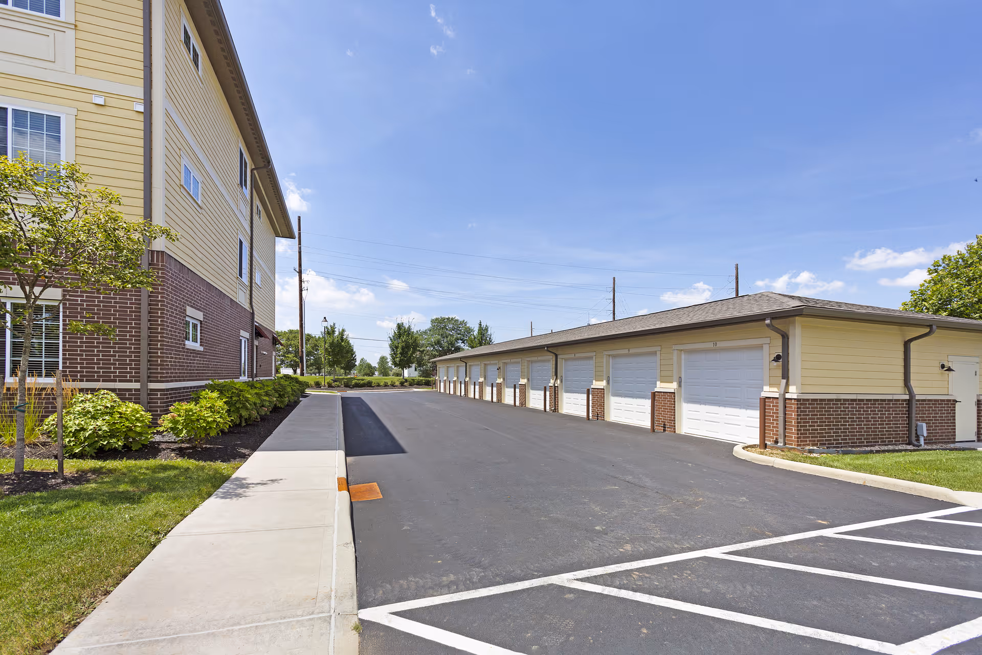 Exterior view of a senior living facility showing a paved driveway with multiple garage doors on the right and a three-story building with yellow siding and brick on the left, under a clear blue sky.