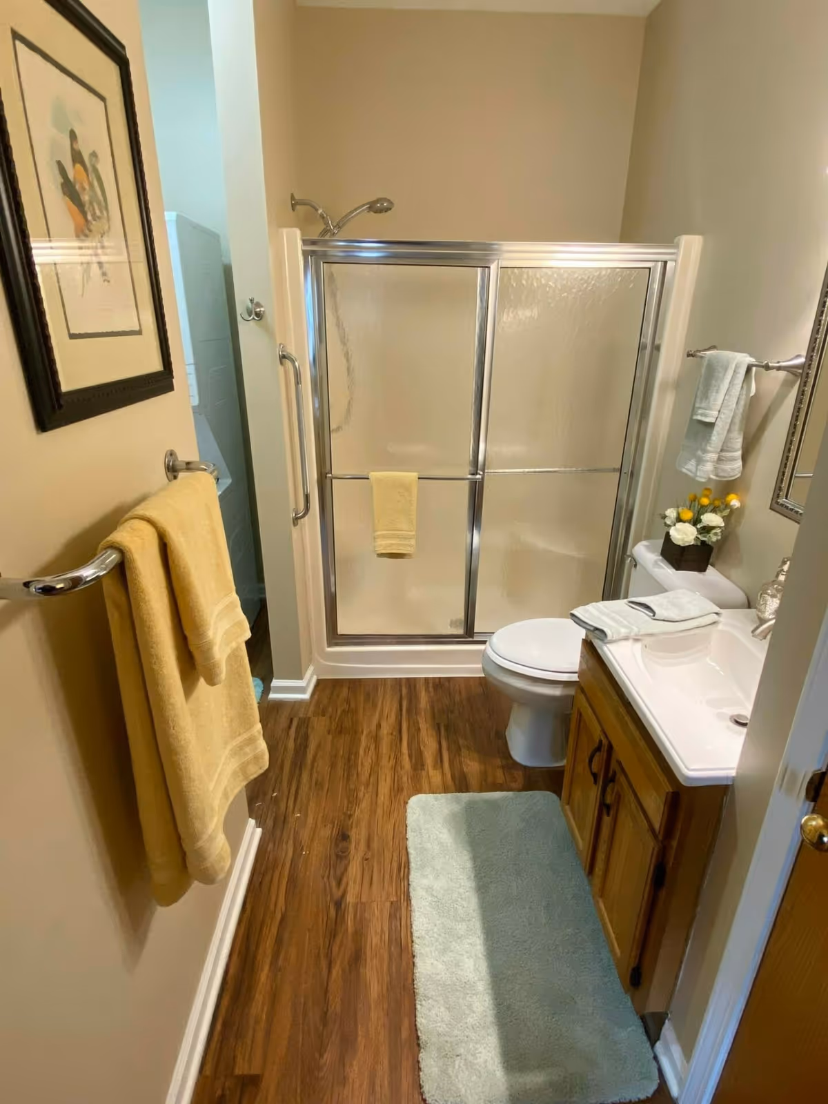 A small bathroom with wood-look flooring, a shower with sliding glass doors, a toilet, and a wooden vanity with a white sink. Yellow and light blue towels hang on towel racks, and a small flower arrangement is placed on the toilet tank. A framed picture is on the wall to the left.