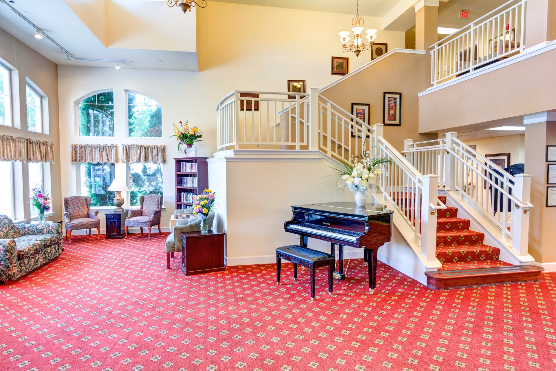 A spacious and well-lit common area with large windows, floral-patterned sofa, two armchairs, a side table with a lamp, a bookshelf, and a grand piano with a bench. The room features a red carpet with a repeating pattern, cream-colored walls, and a staircase with white railings leading to an upper level. There are framed pictures on the walls and flower arrangements on tables and the piano.