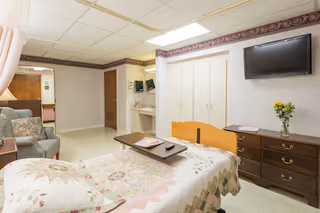 A well-lit patient room in a healthcare facility featuring a bed with a floral quilt, a wooden dresser with a vase of yellow flowers, a wall-mounted flat-screen TV, a comfortable armchair, and a small vanity area with a mirror. The room has light-colored walls with a decorative border near the ceiling and a door leading to another area.