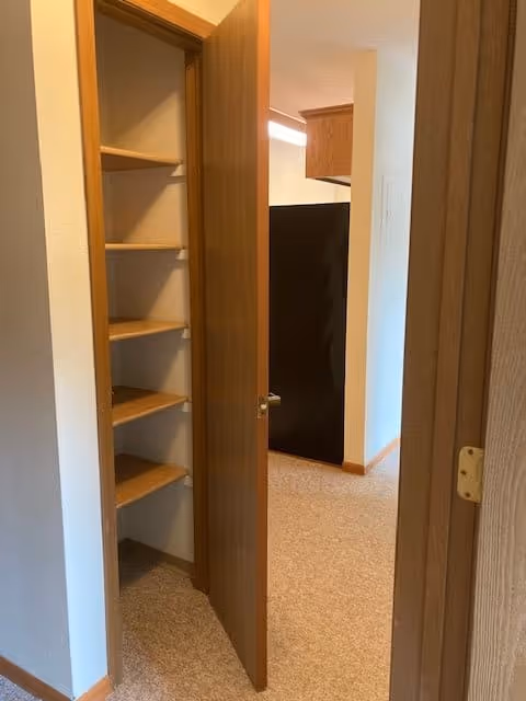 Interior view of a hallway in a senior living facility showing an open wooden door revealing built-in wooden shelves. The hallway carpet is beige, and a black refrigerator is visible in the adjacent kitchen area.