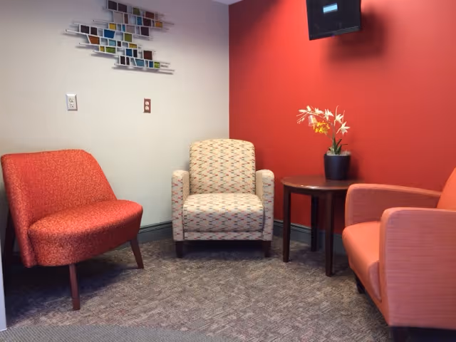 A small seating area with three upholstered chairs arranged around a round wooden side table with a potted plant on it. The walls are painted red and light gray, with a colorful abstract wall decoration above one chair. The floor is carpeted.