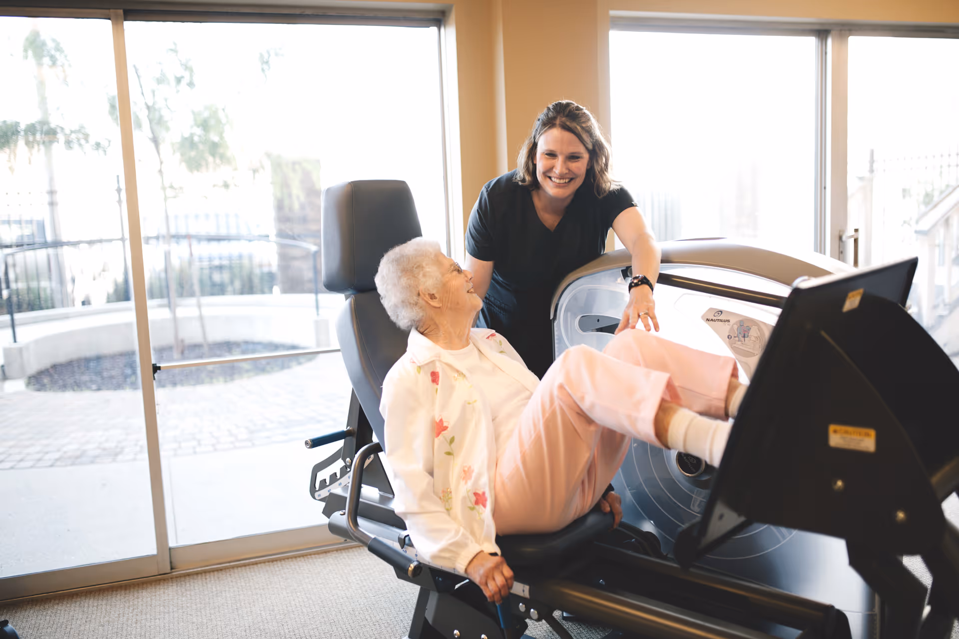 An elderly woman is using a leg press exercise machine indoors while a smiling caregiver or trainer stands beside her, offering support. Large windows in the background let in natural light and show an outdoor patio area.